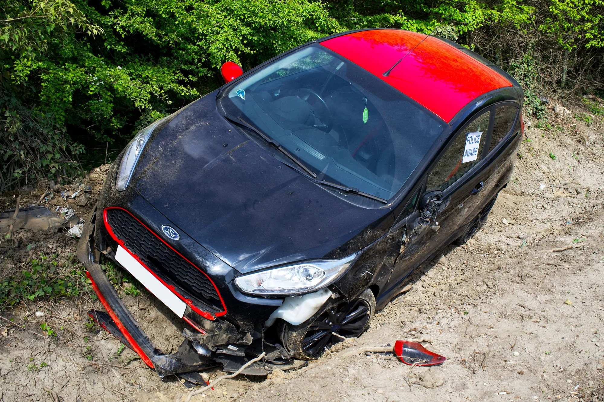 Black hatchback with a red roof sitting nose-down in a ditch, its front bumper torn loose